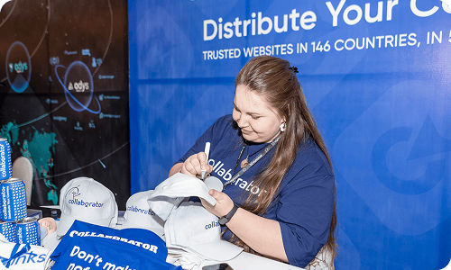 Miembro del equipo de Collaborator con una camiseta de marca firmando camisetas promocionales y materiales en un puesto en la Cumbre de SEO Mastery, con el mensaje de distribución de contenido global de Collaborator visible en el fondo