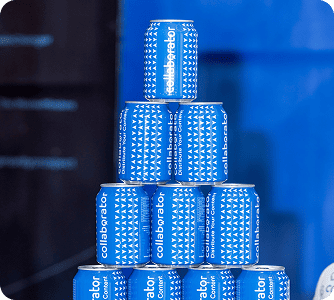 Pila de latas azules de la marca Collaborator dispuestas en múltiples niveles, creando una exhibición llamativa contra un fondo azul